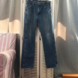 May include: A pair of blue denim jeans hanging on a white wooden hanger against a white wooden shutter background.