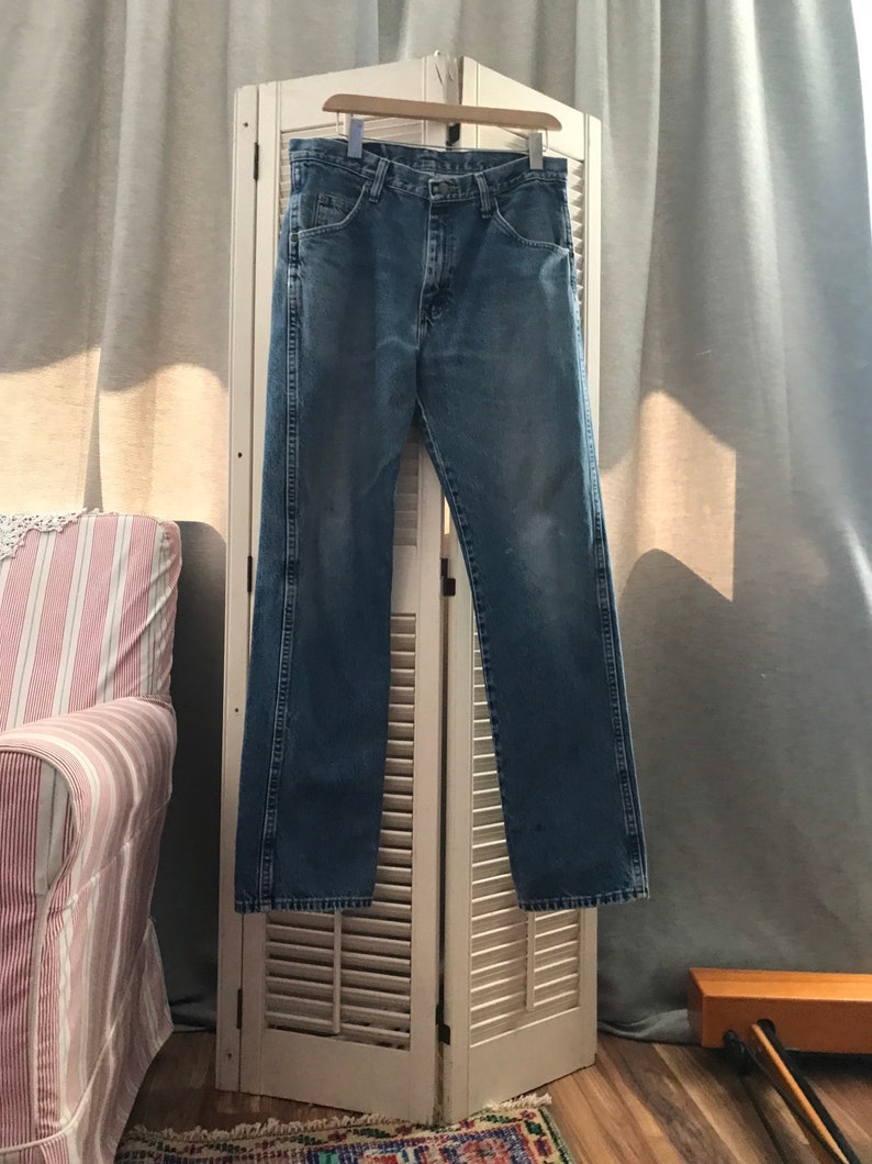 May include: A pair of blue denim jeans hanging on a white wooden hanger against a white wooden shutter background.