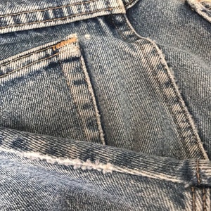 May include: Close-up of the back pocket of a pair of blue denim jeans. The pocket has a light blue denim fabric and brown stitching around the edges.