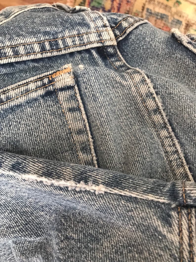May include: Close-up of the back pocket of a pair of blue denim jeans. The pocket has a light blue denim fabric and brown stitching around the edges.
