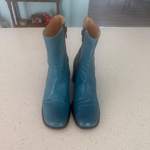May include: A pair of mid-calf boots in a vibrant teal blue leather. These boots feature a square toe design, a block heel, and a vertical zip closure located on the inner side of each shaft. The interior lining is a light tan colour.