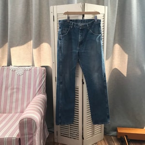 May include: A pair of blue denim jeans hanging on a white wooden folding screen. The jeans have a classic straight leg style and are made of a soft, comfortable fabric.