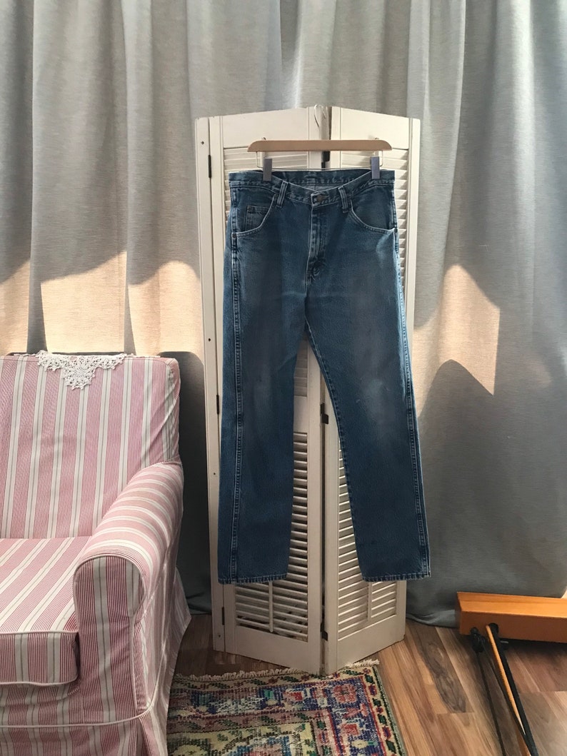 May include: A pair of blue denim jeans hanging on a white wooden folding screen. The jeans have a classic straight leg style and are made of a soft, comfortable fabric.