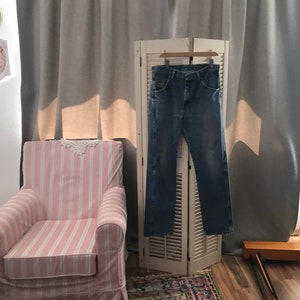 May include: A pair of blue jeans hanging on a white wooden folding screen in front of a pink and white striped armchair. The jeans are hanging on a wooden hanger.
