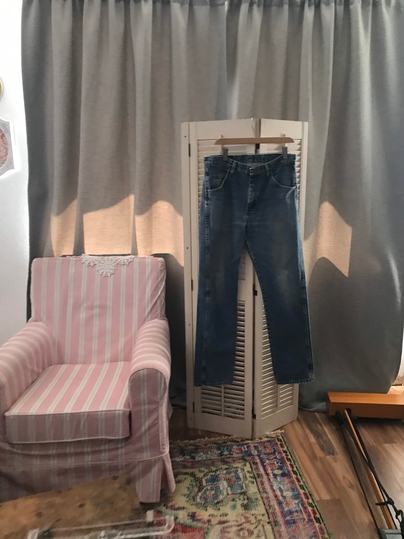 May include: A pair of blue jeans hanging on a white wooden folding screen in front of a pink and white striped armchair. The jeans are hanging on a wooden hanger.