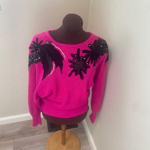 May include: A vibrant pink sweater adorned with black leaf and floral detailing. The sweater is presented on a brown mannequin. The embellishments are highlighted with small white beads. It features long sleeves and a round neckline.