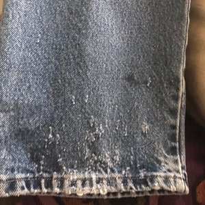 May include: Close-up of the bottom hem of a pair of blue denim jeans. The hem is frayed and worn, showing the white threads underneath.