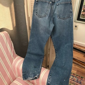 May include: A pair of light blue denim jeans with a faded look. The jeans have a straight leg and a button fly. The back pockets have red stitching detail and a small red label with the word "Wrangler" on it.