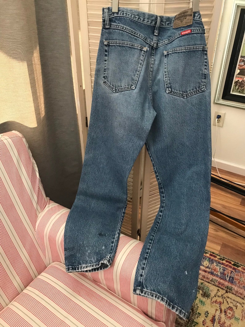 May include: A pair of light blue denim jeans with a faded look. The jeans have a straight leg and a button fly. The back pockets have red stitching detail and a small red label with the word "Wrangler" on it.