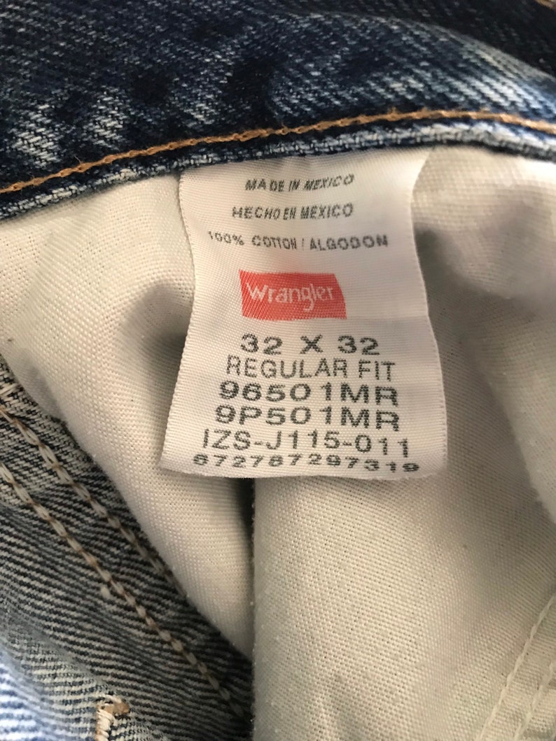May include: A white Wrangler jeans tag with black text. The tag says "Made in Mexico", "Hecho en Mexico", "100% Cotton / Algodon", "Wrangler", "32 X 32", "Regular Fit", "96501MR", "9P501MR", "IZS-J115-011", and "672787297319".