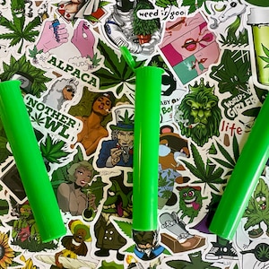 May include: Three bright green, cylindrical tubes with flip-top lids are displayed against a backdrop of cannabis-themed stickers. The stickers feature various illustrations, including cannabis leaves, cartoon characters, and text like "weed is good" and "Nother Bowl".