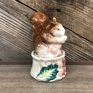 May include: A ceramic squirrel ornament with a brown tail and fur, white belly, and a white base decorated with green leaves and pink berries. The squirrel is perched on the base, holding its paws together. The ornament is on a wooden surface.
