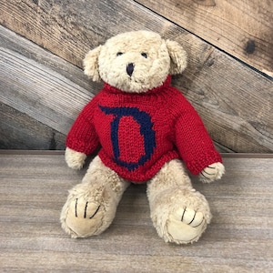 Op de afbeelding: Een pluche teddybeer die een rode gebreide trui draagt met een blauwe letter "D" op de voorkant. De beer zit op een houten oppervlak.