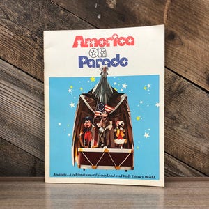 May include: A vintage brochure for the "America on Parade" show at Disneyland and Walt Disney World. The cover features a cartoon eagle with Mickey Mouse, Donald Duck, Goofy, and Pluto on a drum.