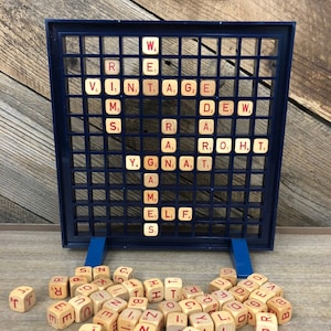 May include: A blue plastic game board with a grid of squares, each containing a letter tile. The tiles are made of wood and have red letters on them. There are also a number of loose letter tiles scattered around the board.