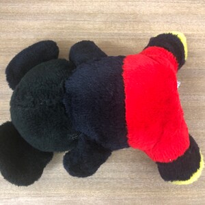 以下が含まれることがあります： 黒と赤のぬいぐるみで、黄色のアクセントがあります。おもちゃは背中を上にして、足を上げています。