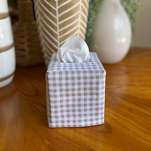 May include: A square tissue box cover with a brown and white gingham pattern. The box is filled with white tissues and sits on a wooden surface. Decorative vases are in the background.