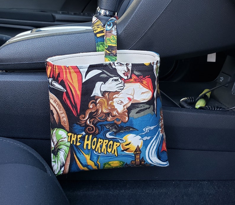 Op de afbeelding: Een stoffen auto-afvalzak met een vintage horrorfilmafbeelding. De tas heeft een kleurrijke print met afbeeldingen van vampiers, vleermuizen en een spookkasteel. De tekst "THE HORROR" staat erop.