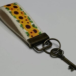 May include: A key fob with a sunflower pattern on a cream-colored background. The key fob is attached to a bronze-colored metal key and hardware. The key is a vintage style.