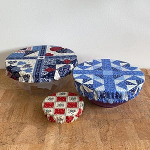 May include: Three round fabric bowl covers with elastic edges. The largest cover features a blue, white, and red patchwork pattern. The medium cover has a blue and white geometric quilt design. The smallest cover displays a red and white floral grid.