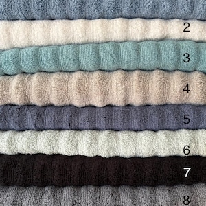 Op de afbeelding: Een stapel van acht gevouwen handdoeken met een geribbelde textuur in verschillende kleuren, genummerd van &eacute;&eacute;n tot acht. De kleuren omvatten tinten blauw, cr&egrave;me, groenblauw, beige, donkergrijs, lichtgroen, donkerbruin en antracietgrijs.