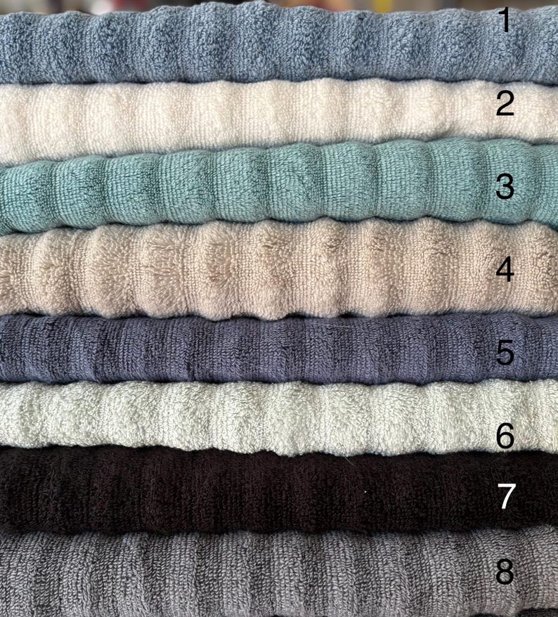 Op de afbeelding: Een stapel van acht gevouwen handdoeken met een geribbelde textuur in verschillende kleuren, genummerd van &eacute;&eacute;n tot acht. De kleuren omvatten tinten blauw, cr&egrave;me, groenblauw, beige, donkergrijs, lichtgroen, donkerbruin en antracietgrijs.