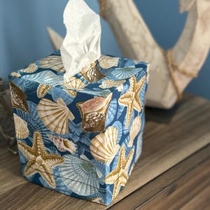 May include: A square tissue box cover with a blue background and a pattern of seashells and starfish in shades of white, tan, and brown. White tissues are pulled from the top opening. The box is sitting on a wooden surface.