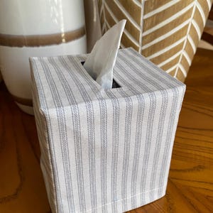 May include: A square tissue box cover with a light gray and white striped pattern. A white tissue is pulled from the top opening. The box is on a wooden surface, with decorative ceramic vases in the background.