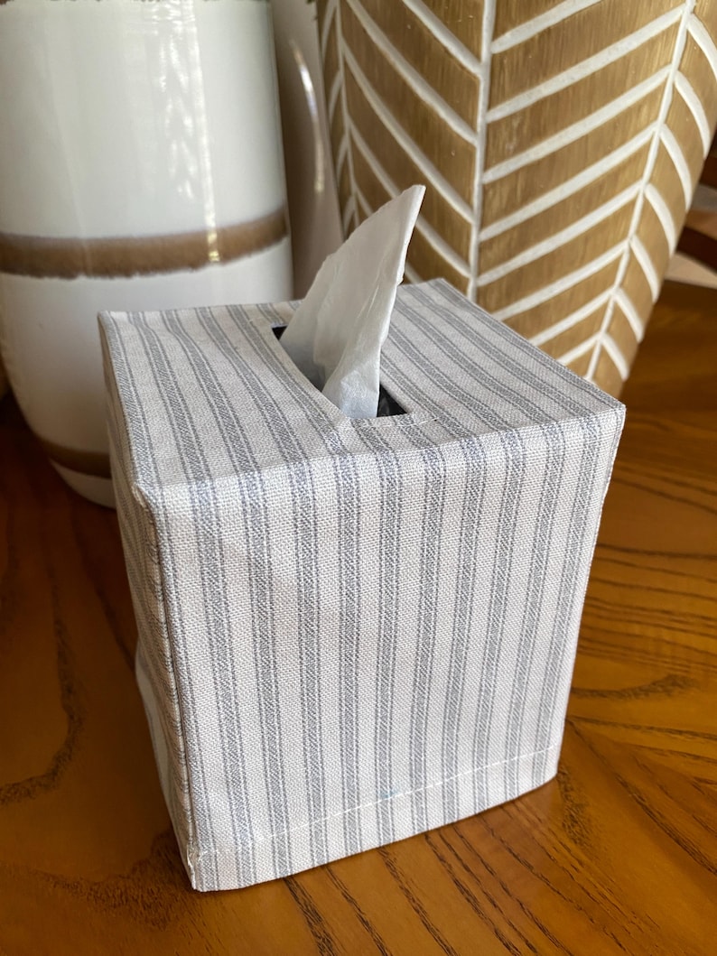 May include: A square tissue box cover with a light gray and white striped pattern. A white tissue is pulled from the top opening. The box is on a wooden surface, with decorative ceramic vases in the background.