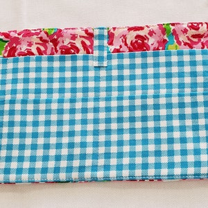 May include: A rectangular fabric item with a blue and white gingham pattern on the front. The top section features a floral print with pink roses and green leaves. A small fabric loop and tab closure are visible.