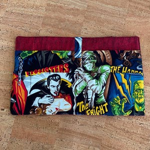 Op de afbeelding: Een rechthoekige stoffen portemonnee met een kleurrijke collage van klassieke filmmonsters. Het ontwerp bevat Dracula, de mummie en Frankenstein's monster, met de tekst "The Fright" en "The Horrors". De boven- en achterkant zijn effen rode stof.