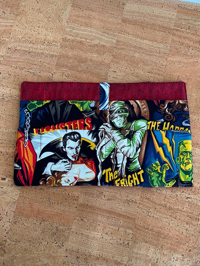 Op de afbeelding: Een rechthoekige stoffen portemonnee met een kleurrijke collage van klassieke filmmonsters. Het ontwerp bevat Dracula, de mummie en Frankenstein's monster, met de tekst "The Fright" en "The Horrors". De boven- en achterkant zijn effen rode stof.