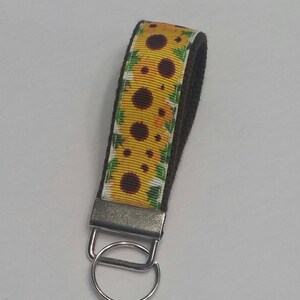 May include: A sunflower-themed keychain wristlet. The wristlet features a yellow ribbon with a sunflower pattern and a dark brown backing. It has a silver-colored metal clasp and a key ring.