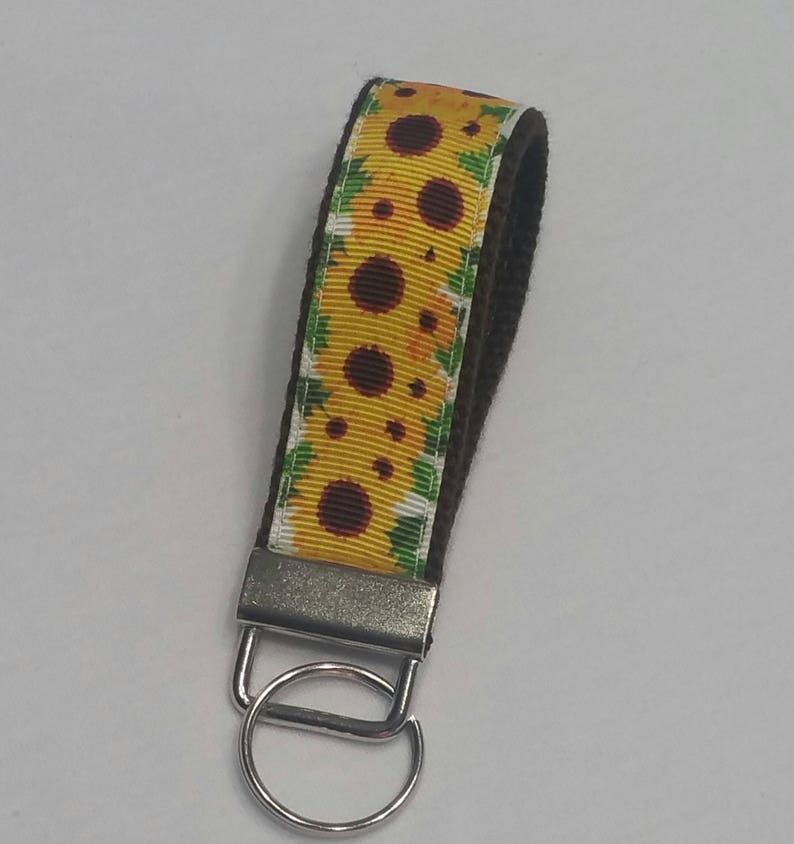 May include: A sunflower-themed keychain wristlet. The wristlet features a yellow ribbon with a sunflower pattern and a dark brown backing. It has a silver-colored metal clasp and a key ring.