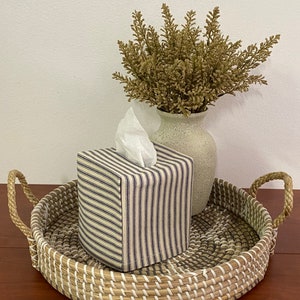 May include: A square tissue box cover with a blue and white striped pattern sits on a woven tray with handles. A vase with dried foliage is next to the tissue box. The tray is made of natural fibers.