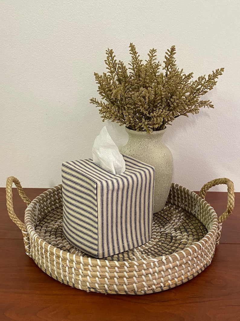 May include: A square tissue box cover with a blue and white striped pattern sits on a woven tray with handles. A vase with dried foliage is next to the tissue box. The tray is made of natural fibers.
