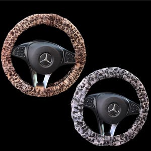May include: Two steering wheel covers with a leopard print design. One cover is brown and tan, the other is gray and white. The covers are circular and fit over a black steering wheel with a silver Mercedes-Benz logo.