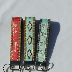 May include: Three fabric keychains with metal key rings. One is red with white arrows, another is turquoise with gold geometric shapes, and the third is gray with white owl designs. The keychains have a woven texture.