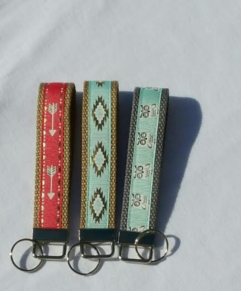 May include: Three fabric keychains with metal key rings. One is red with white arrows, another is turquoise with gold geometric shapes, and the third is gray with white owl designs. The keychains have a woven texture.