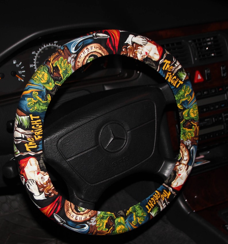Op de afbeelding: Een zwart stuurwiel met een kleurrijke hoes met vintage horrorfilm illustraties. De hoes toont afbeeldingen van monsters, vrouwen en de tekst "The Fright" in een herhalend patroon. Het dashboard van de auto is zichtbaar op de achtergrond.