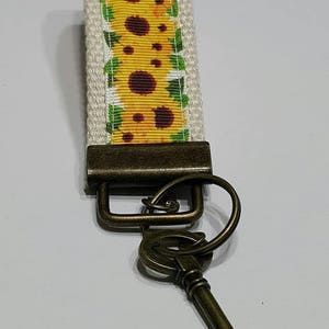 May include: A key fob with a sunflower design. The key fob has a cream-colored background with a yellow sunflower pattern. The hardware is a dark bronze color, and a bronze-colored key is attached.