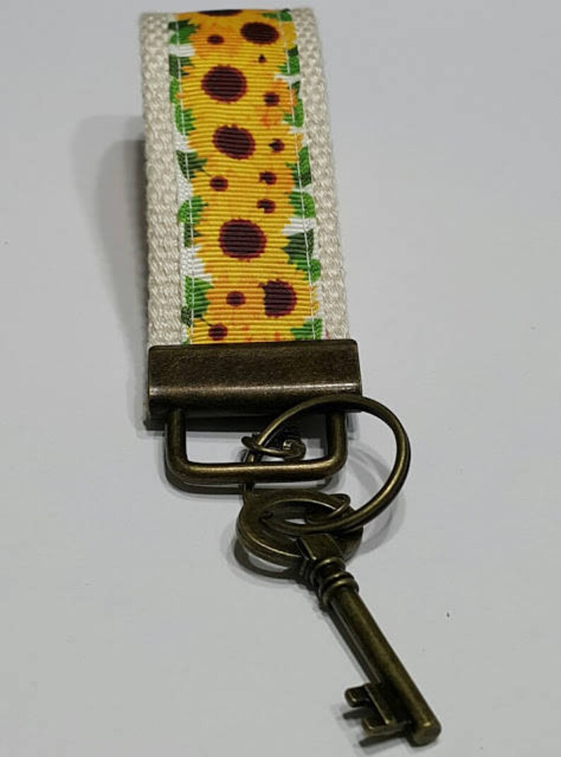 May include: A key fob with a sunflower design. The key fob has a cream-colored background with a yellow sunflower pattern. The hardware is a dark bronze color, and a bronze-colored key is attached.