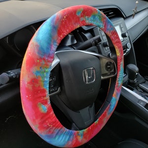May include: A colorful steering wheel cover with a vibrant tie-dye pattern in shades of red, blue, and yellow. The cover is wrapped around a black steering wheel, adding a pop of color to the car's interior. The cover is designed to fit a standard steering wheel.