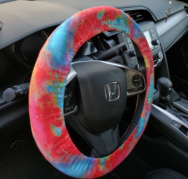 May include: A colorful steering wheel cover with a vibrant tie-dye pattern in shades of red, blue, and yellow. The cover is wrapped around a black steering wheel, adding a pop of color to the car's interior. The cover is designed to fit a standard steering wheel.