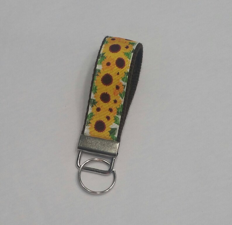 May include: A sunflower patterned wristlet keychain. The yellow ribbon features brown sunflowers and green leaves. The wristlet is attached to a silver metal clasp and key ring. The wristlet is approximately 6 inches long.