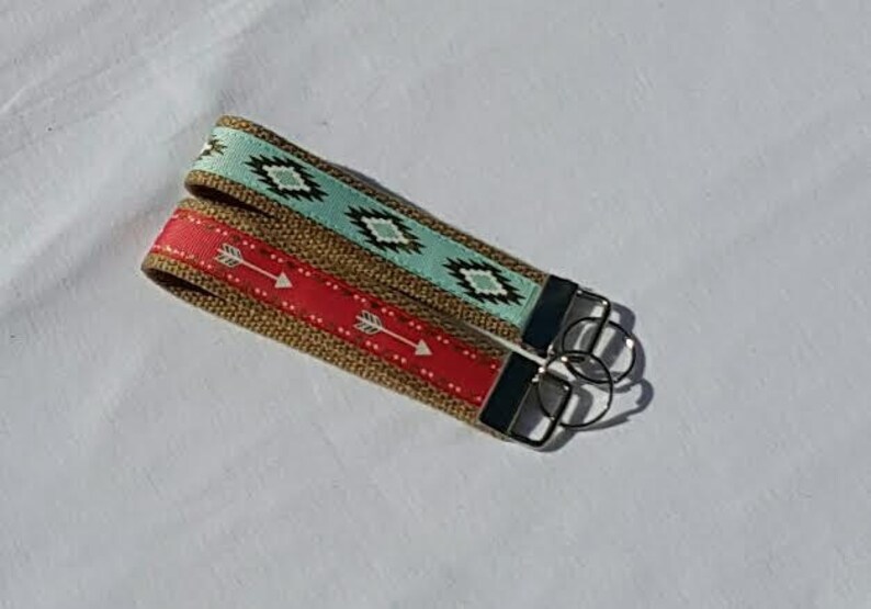 May include: Two keychains with woven burlap straps. One has a red strap with white arrows, the other has a turquoise strap with a geometric pattern. Both have silver key rings and black end pieces.