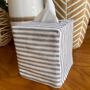 May include: A square tissue box cover with a black and white striped pattern. The cover is made of fabric and has a simple, clean design. White tissues are visible at the top of the box. The box is sitting on a wooden surface.