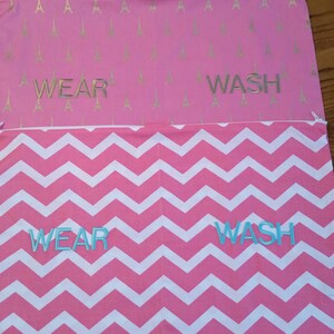 May include: Two pink fabric bags with zippers. The top bag has a gold Eiffel Tower pattern and the word "WEAR" and "WASH" in silver. The bottom bag has a white and pink chevron pattern with the words "WEAR" and "WASH" in blue.