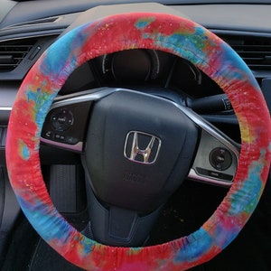 May include: A colorful steering wheel cover with a vibrant tie-dye pattern in shades of red, blue, and yellow. The cover is wrapped around a black steering wheel, enhancing the car's interior with a splash of color.