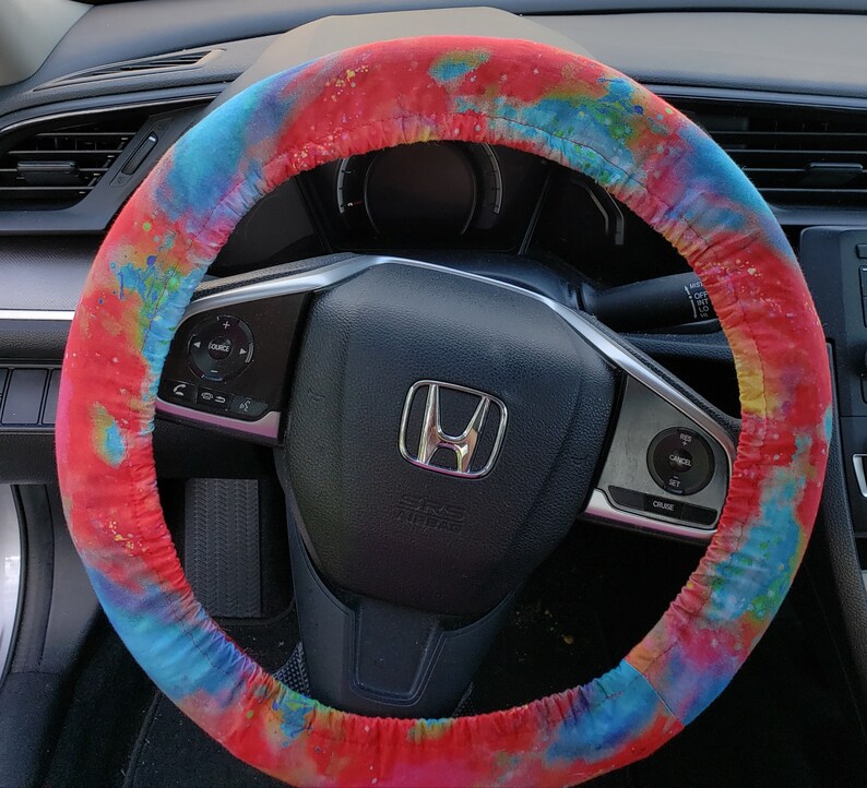 May include: A colorful steering wheel cover with a vibrant tie-dye pattern in shades of red, blue, and yellow. The cover is wrapped around a black steering wheel, enhancing the car's interior with a splash of color.